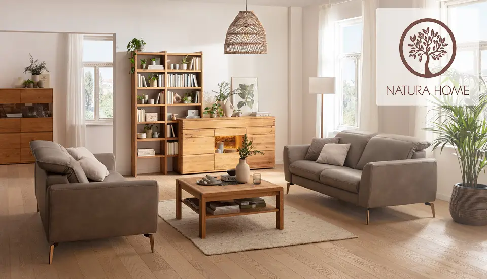Helles Wohnzimmer mit zwei grauen Sofas, Holzmöbeln, Bücherregalen und Pflanzen, natürliche Lichtverhältnisse durch große Fenster, Markenlogo „NATURA HOME“ oben rechts.